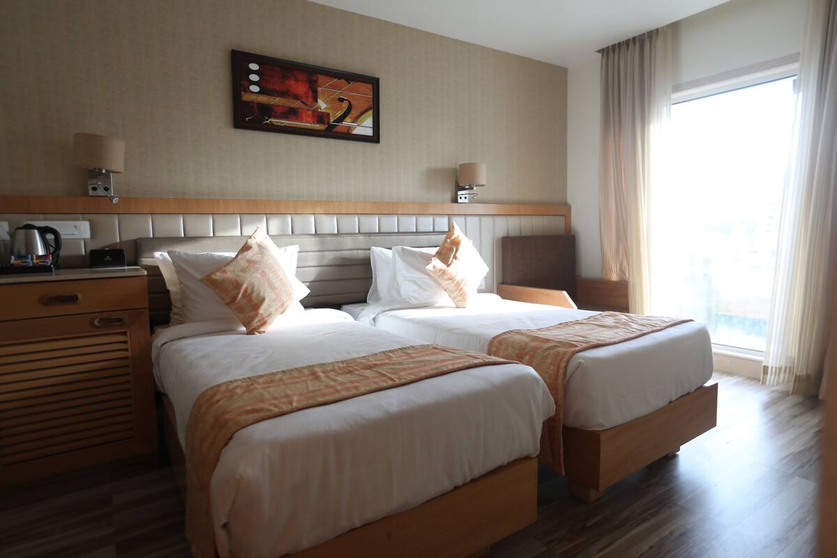 A hotel room with two beds, a headboard, a desk with a lamp, a painting on the wall, and a window with curtains.