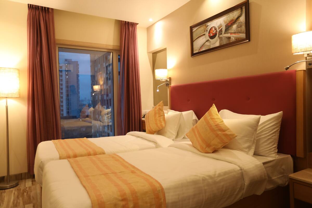 A hotel room with a large bed, two pillows, a red headboard, a nightstand, a lamp, and a window with curtains.