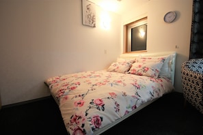 A bedroom with a floral bedspread, a bedside table, a chair, a wall clock, and a window with blinds.