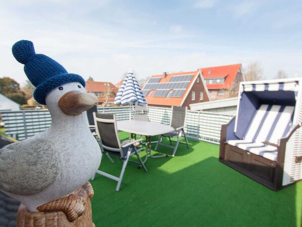 A decorative duck statue on a wooden post, a table with chairs, and a striped umbrella in a patio area with green artificial turf and red-roofed buildings in the background.