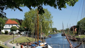 A riverside town with a white building with a red roof, a dock with boats, and a statue.