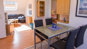 A modern living room with a glass dining table, a sofa, and a TV. There is a wooden cabinet and a small shelf with decorative items.