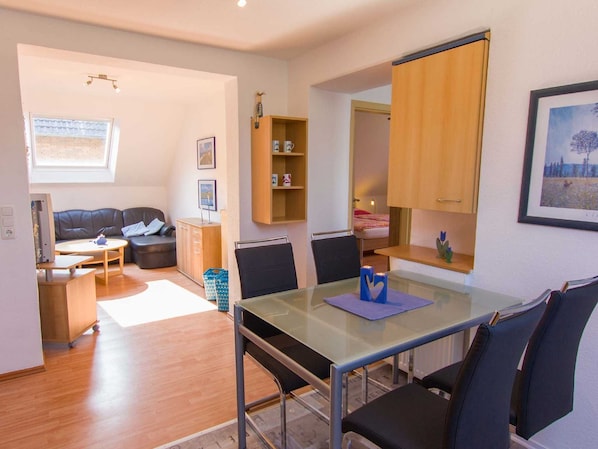A modern living room with a glass dining table, a sofa, and a TV. There is a wooden cabinet and a small shelf with decorative items.