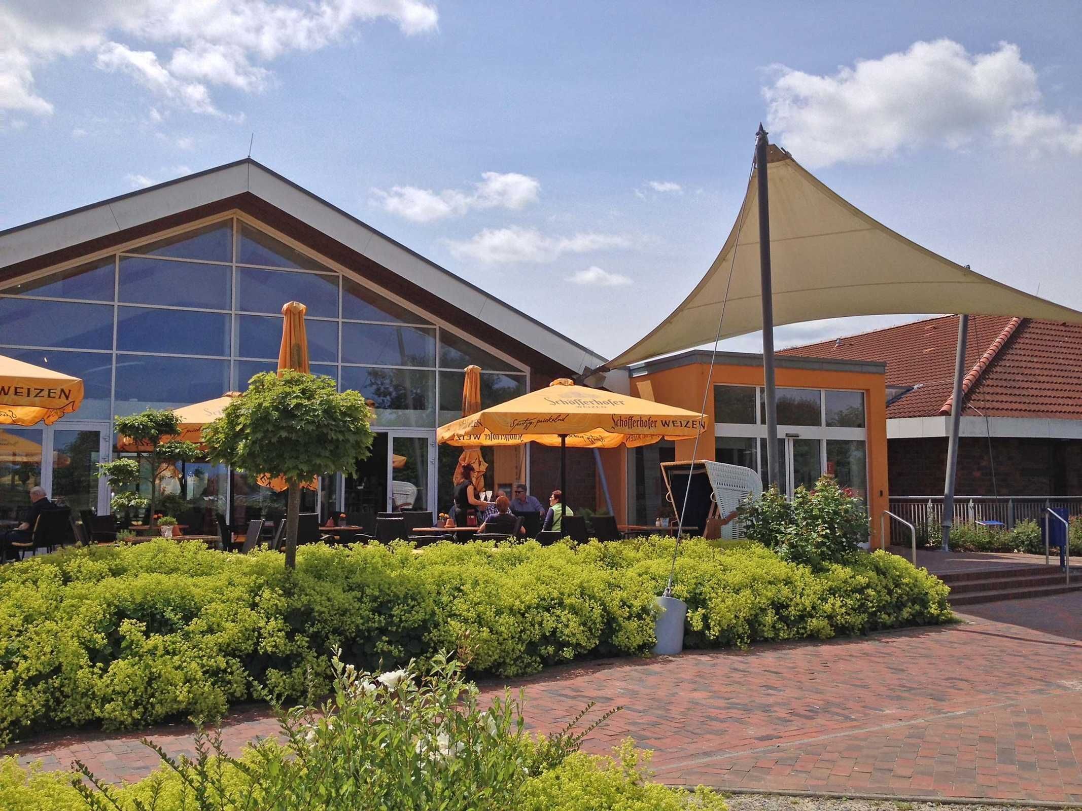 Un espace extérieur aménagé avec des tables et des chaises sous des parasols jaunes, un bâtiment doté de grandes fenêtres et un jardin paysager.