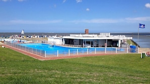 A swimming pool area with a building in the background, a grassy field, and a clear sky.