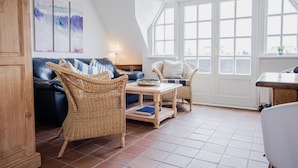 Ein gemütliches Wohnzimmer mit einem dunkelblauen Sofa, Korbstühlen, einem Holztisch und einem großen Fenster mit weißen Fensterläden.