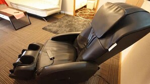 Un fauteuil de massage doté d’un repose-pieds et d’un porte-gobelet, placé dans une pièce dont le sol est recouvert d’une moquette et où l’on aperçoit un lit au fond.