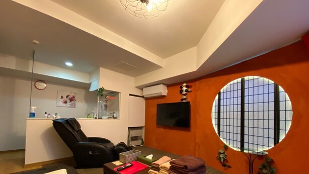 Une chambre moderne équipée d’un fauteuil de massage, d’une petite table avec des serviettes, d’un téléviseur et d’une fenêtre à motifs géométriques.