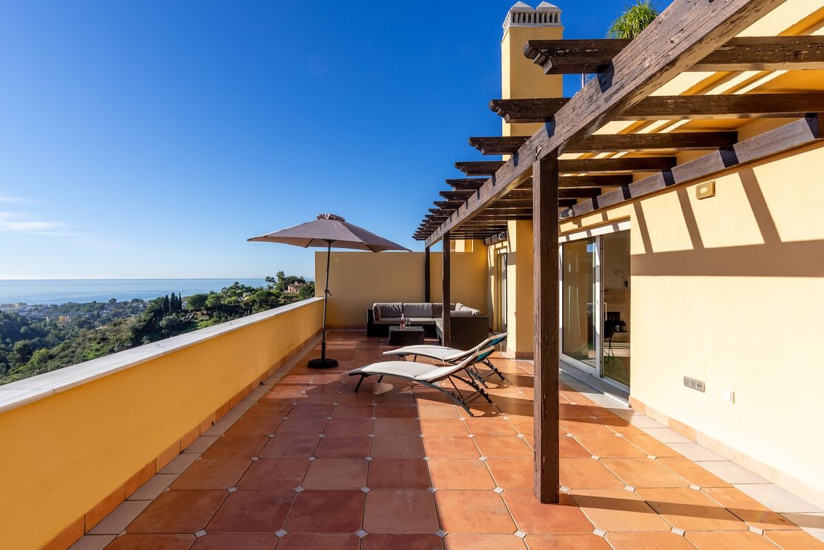 A balcony with a view of the sea, a sofa, two lounge chairs, and a pergola structure.