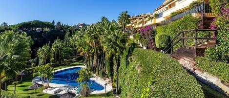 Una piscina rodeada de abundante vegetación y palmeras, con un edificio que cuenta con balcones y flores moradas.