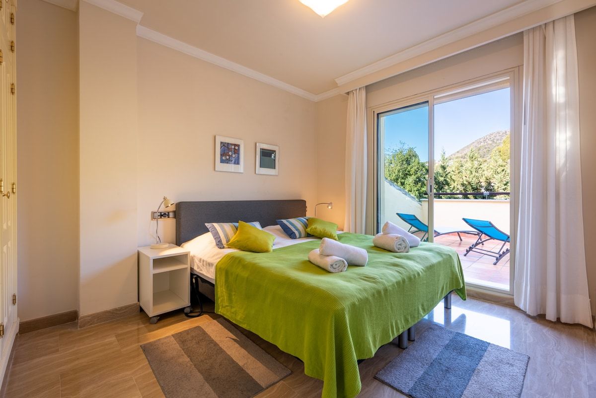 A hotel room with a bed, a nightstand, a desk, a chair, and a view of the outdoors.