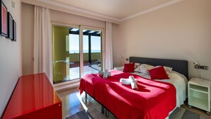 Une chambre d’hôtel comprenant un lit, une table de chevet, un bureau, une chaise, une porte-fenêtre coulissante donnant sur un balcon et un banc rouge.