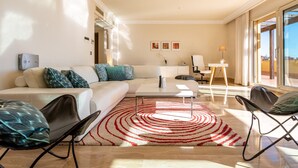 Un salon moderne avec un canapé, une table basse et des fauteuils. Une grande fenêtre offre une vue sur l’extérieur.