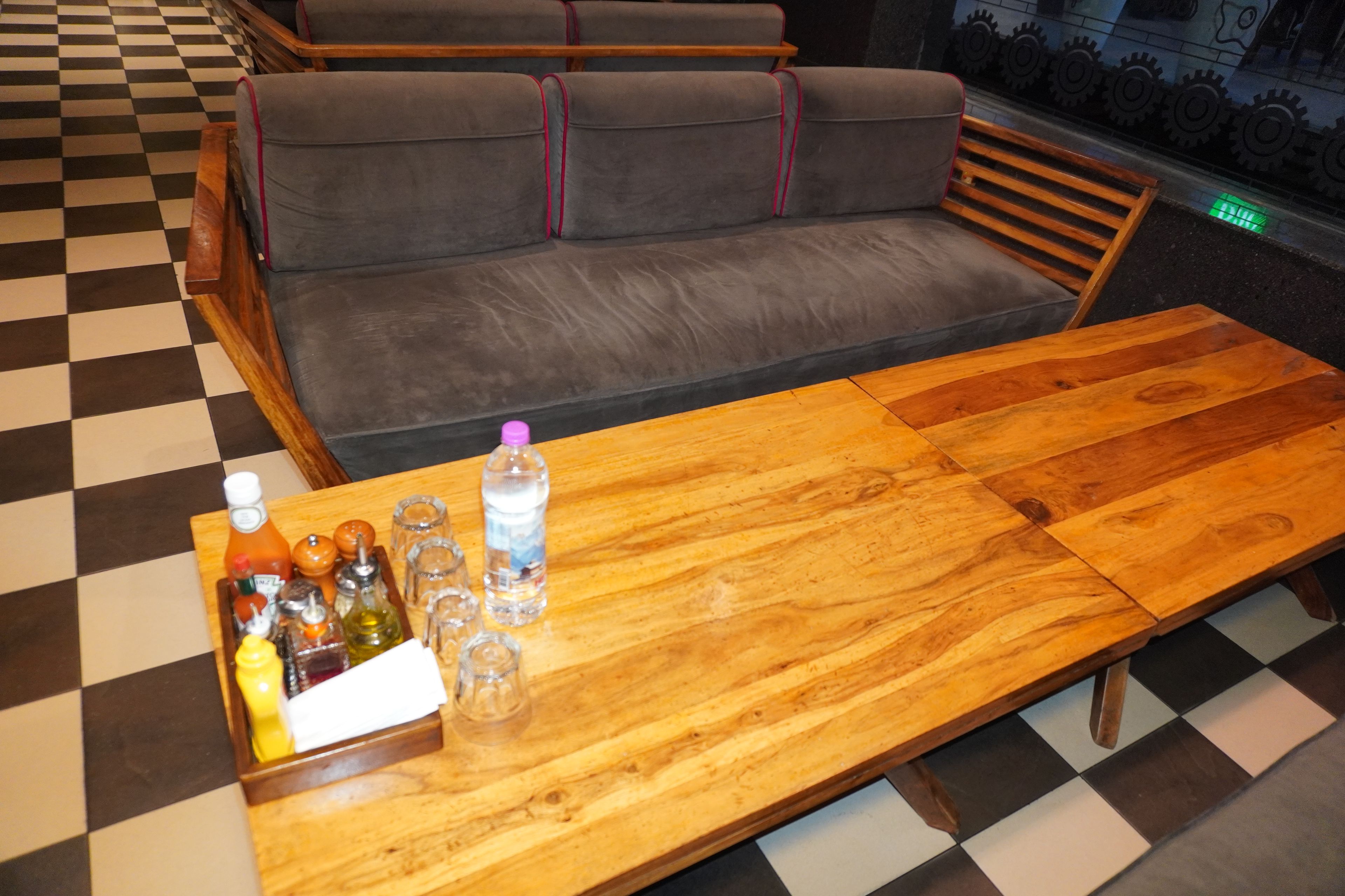 A checkered floor, a wooden table with a bottle of water, and a grey sofa with red piping.