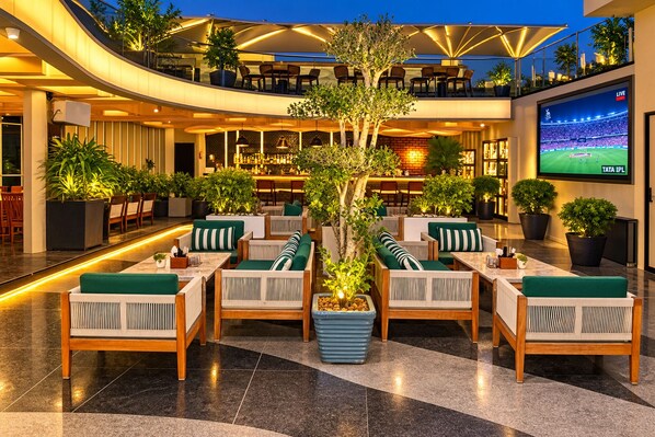 A spacious indoor area with multiple seating arrangements, a bar area, and a large screen displaying a sports event. - Hotel Las Vegas (Jaipur)