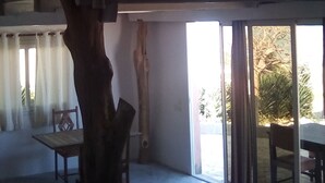 Une pièce spacieuse avec un plafond haut, une grande sculpture en bois représentant un arbre, une table à manger avec des chaises et une porte-fenêtre coulissante donnant sur un espace extérieur agrémenté de plantes.