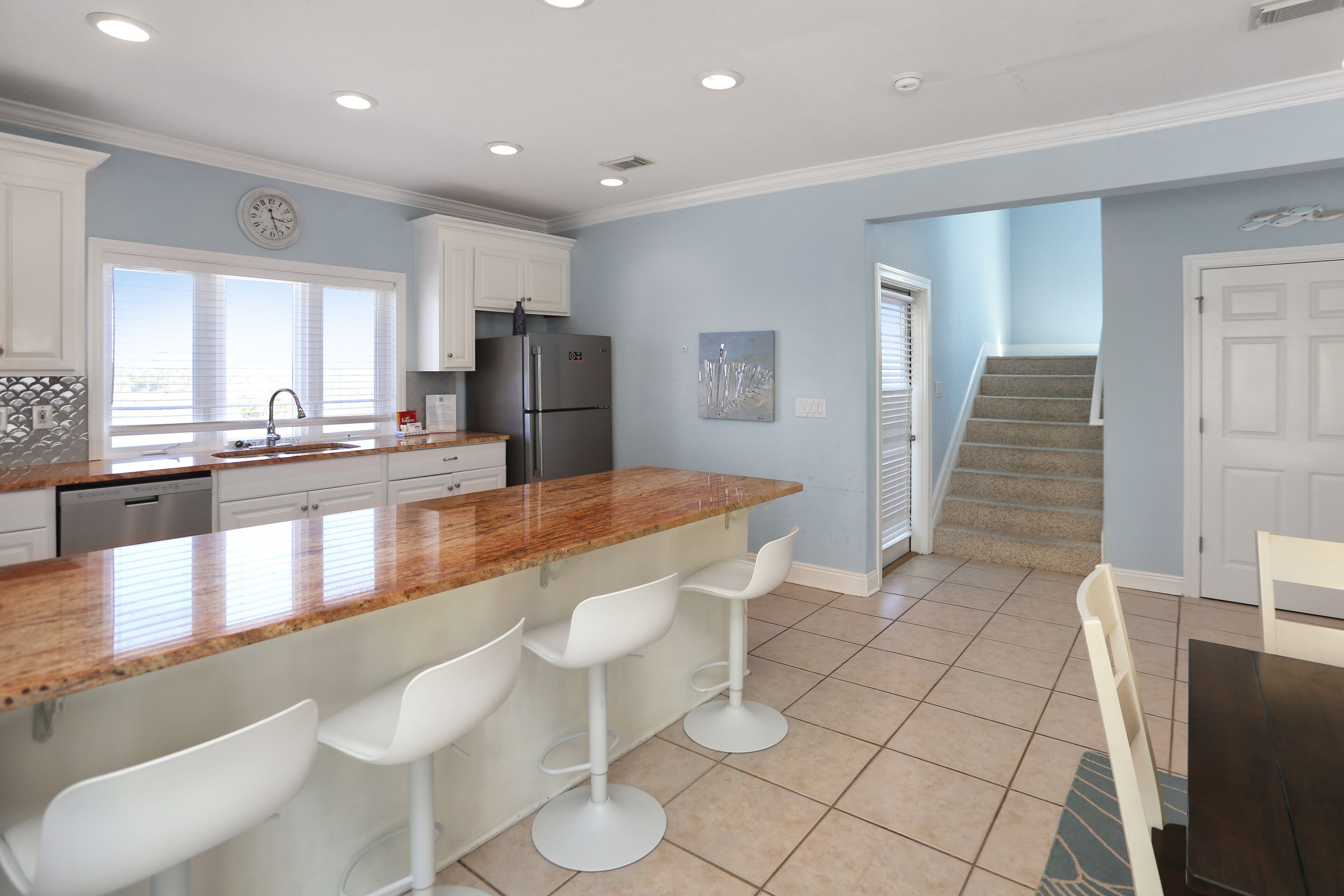 A kitchen with a central island, white bar stools, a stainless steel refrigerator, and a window with blinds.