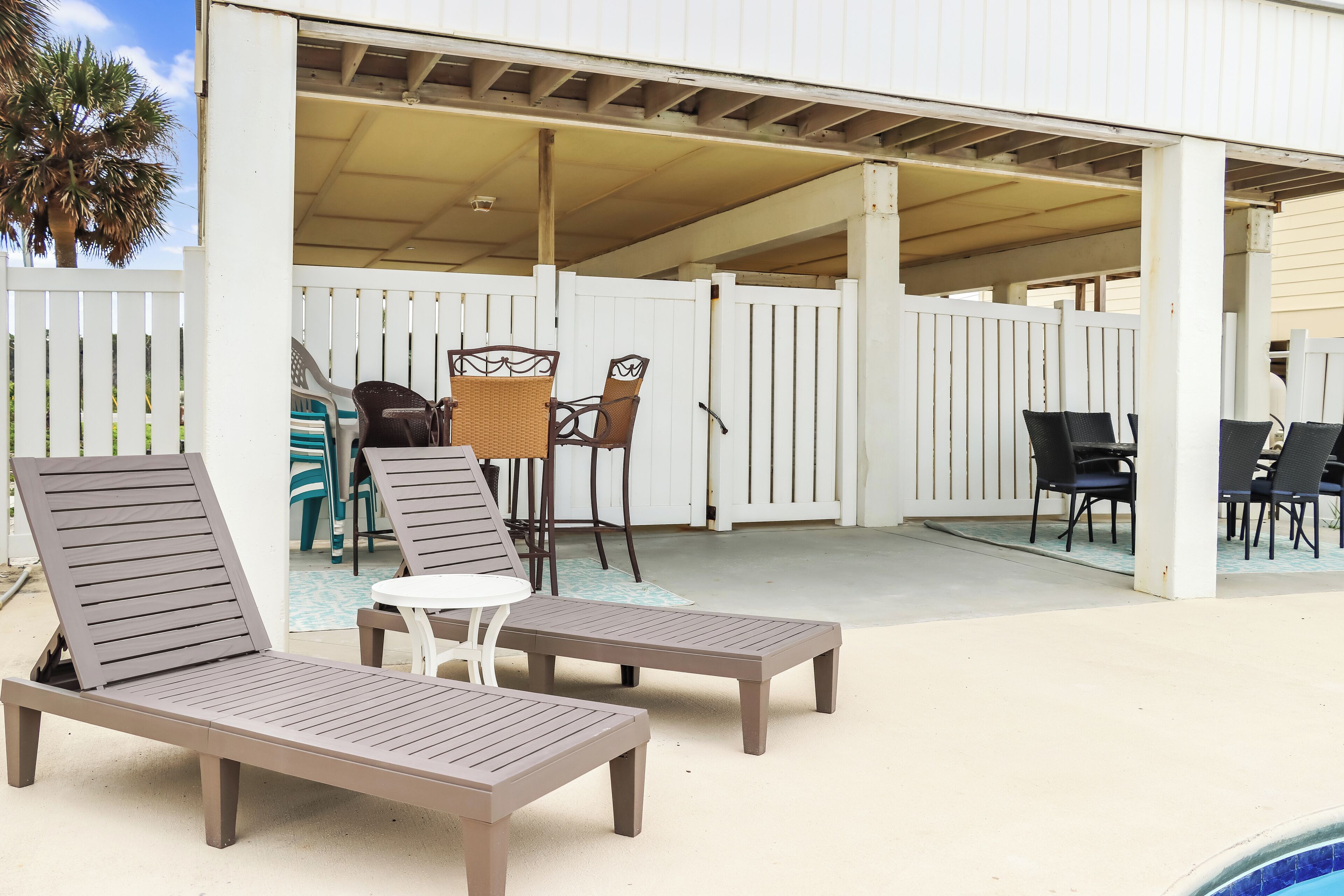 Un espace extérieur aménagé, abrité et doté de chaises longues et d’une table, le tout délimité par une clôture blanche.