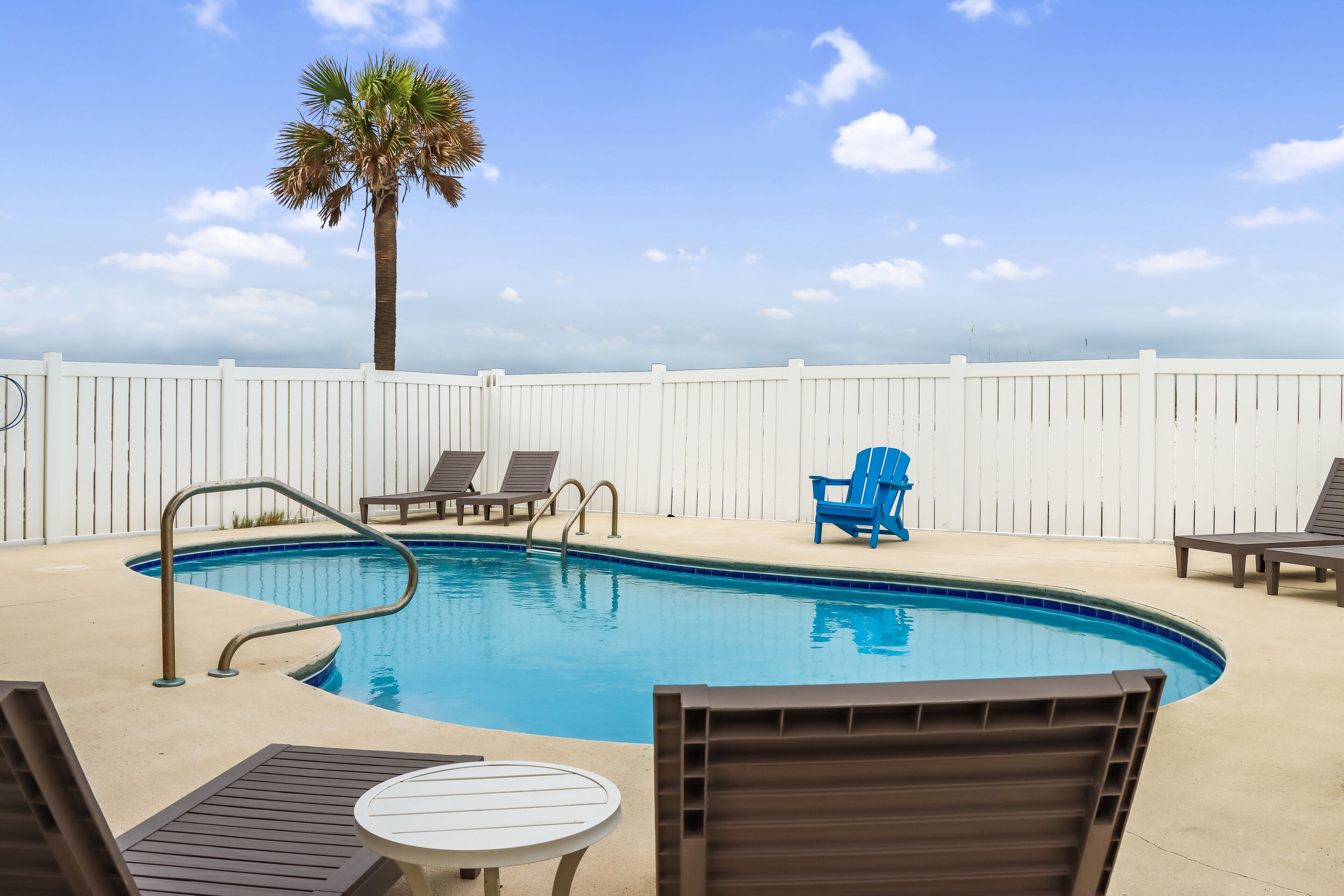 A pool area with a white fence, lounge chairs, and a palm tree.
