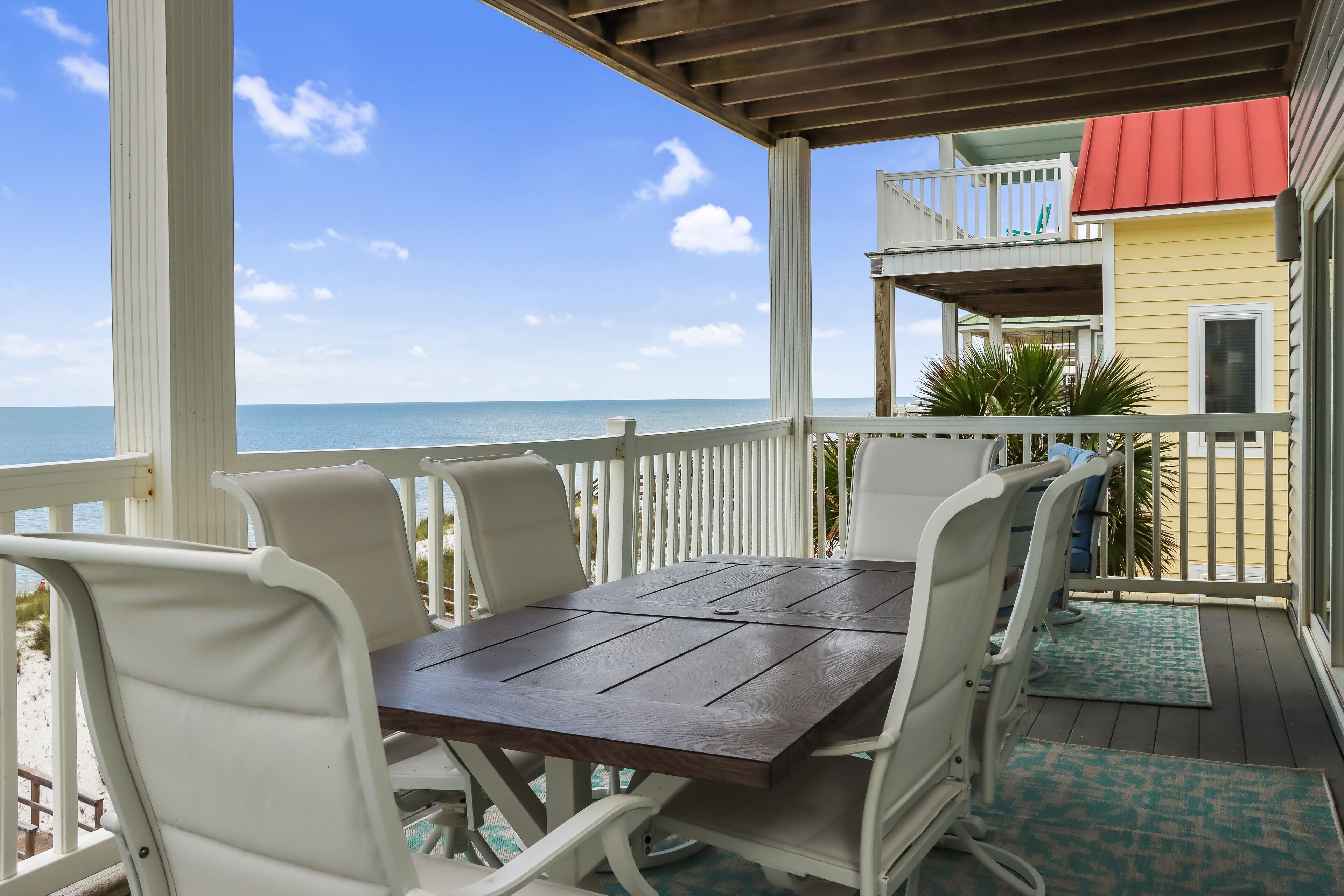A covered outdoor area with a wooden table and white chairs, overlooking a beach and a yellow building with a red roof.