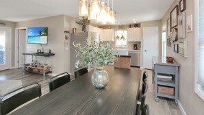 A modern kitchen with a dining area, featuring a large wooden table, hanging lights, and a TV on the wall. - Single Level, Close To Beach, Great Beach View Covered Deck ~ Salt Cured (Cape San Blas)