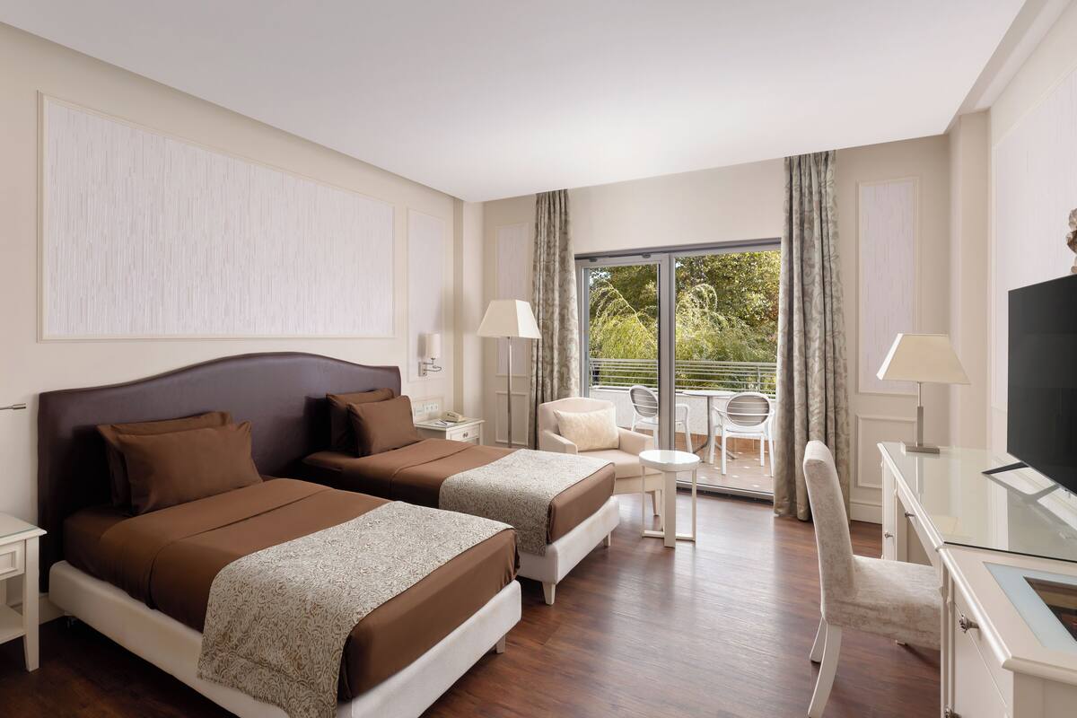 Quarto de hotel com duas camas, uma escrivaninha, uma cadeira, uma televisão e varanda com vista para árvores.