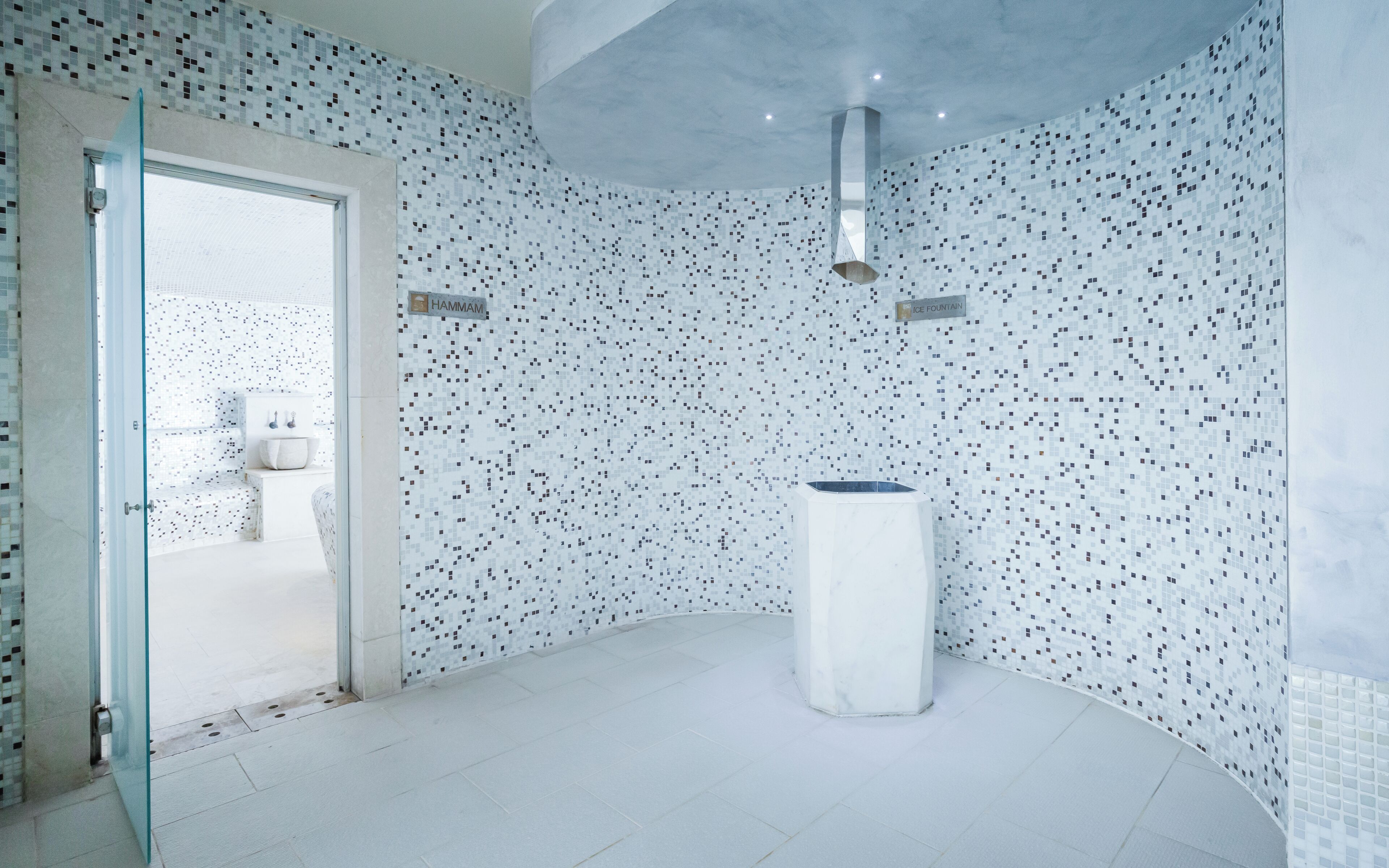 A modern bathroom with a mosaic tile wall, a large white sink, and a glass door leading to a bathroom area.