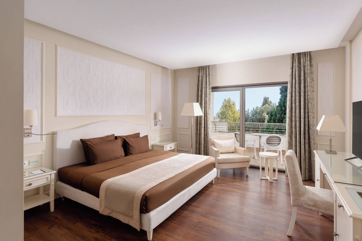 Quarto de hotel com uma cama grande, uma escrivaninha com cadeira, televisão e varanda com vista para árvores.