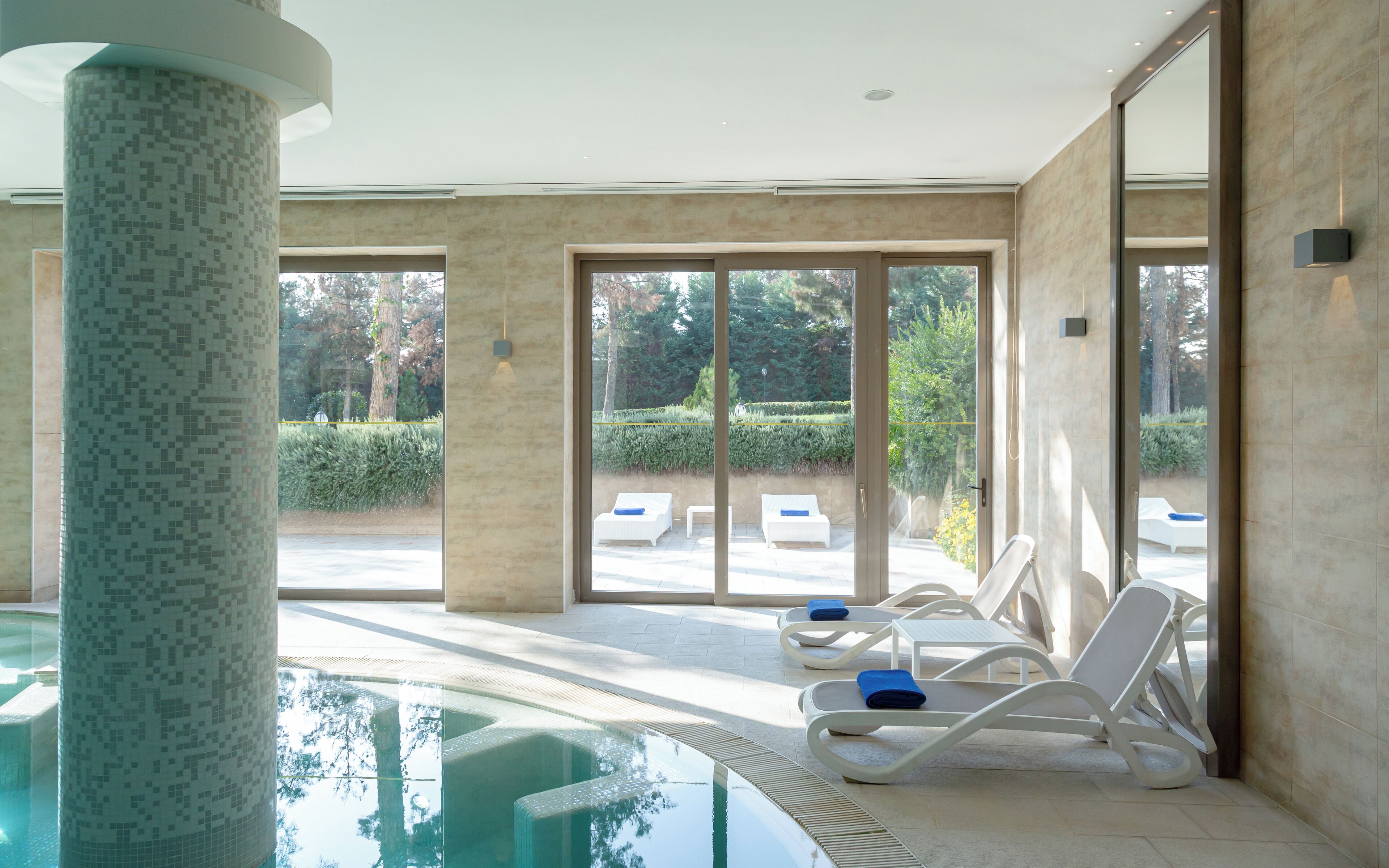 A modern indoor swimming pool area with lounge chairs and a glass wall offering a view of the outdoors.