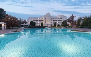A large, clear swimming pool with a tiled deck, surrounded by a modern building with multiple balconies and a well-maintained lawn.