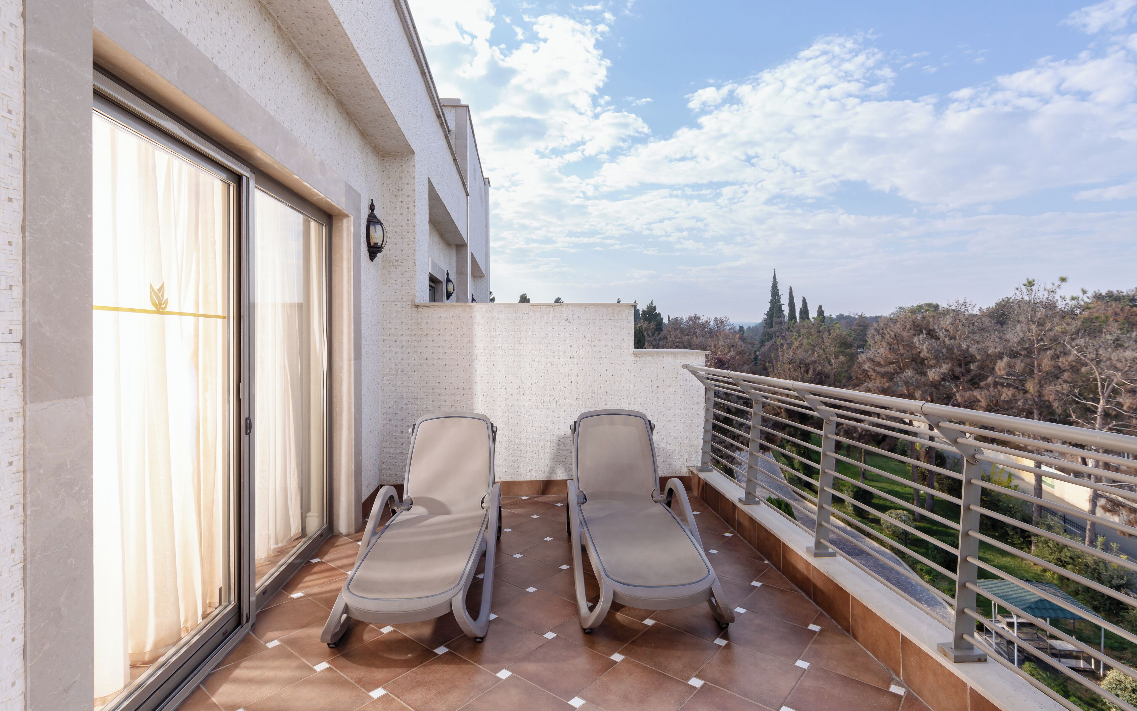 A balcony with two lounge chairs, a large window with curtains, and a view of trees and buildings.