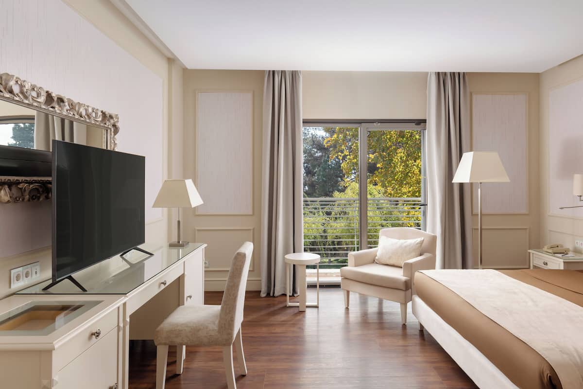 Quarto de hotel com cama, escrivaninha com televisão, duas cadeiras, mesa pequena e janela com cortinas.