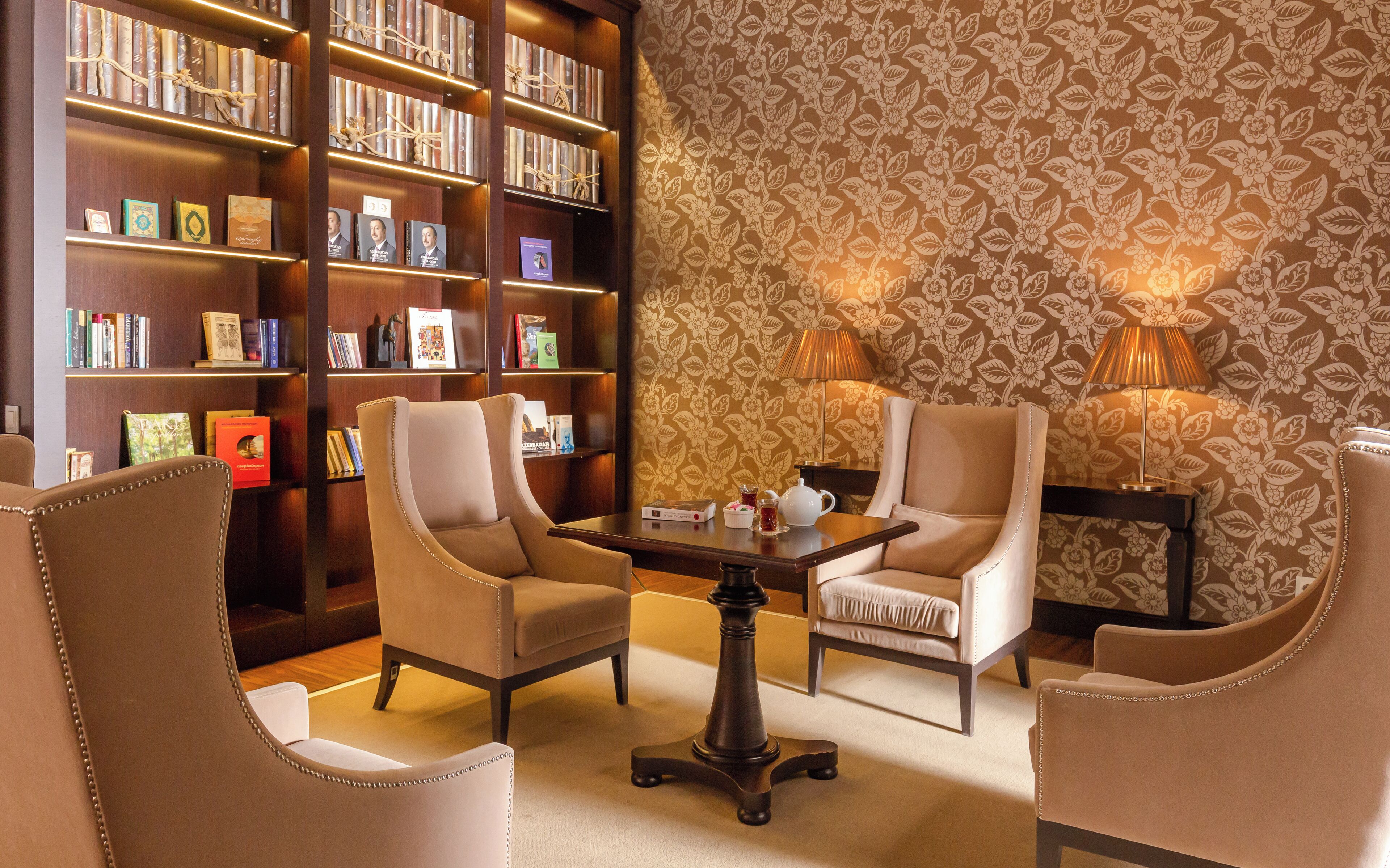 A library with bookshelves, two armchairs, a table with a teapot, and two lamps.