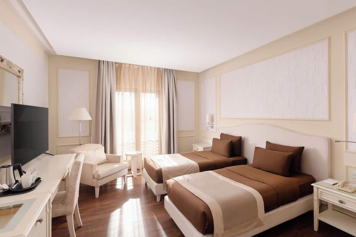 Quarto de hotel com duas camas, uma escrivaninha, uma cadeira, televisão e janela com cortinas.