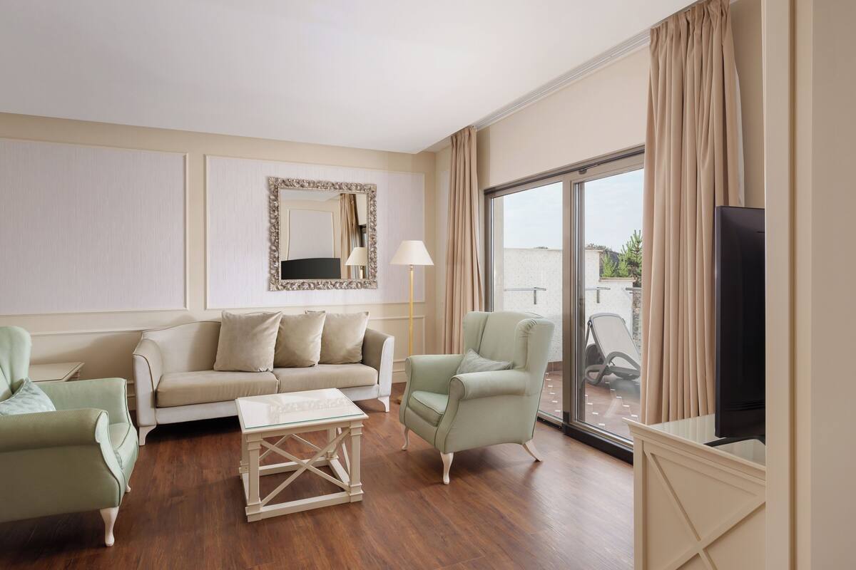 Uma sala de estar com sofá, poltronas, mesa de centro de vidro, televisão de tela plana e uma janela grande com cortinas.