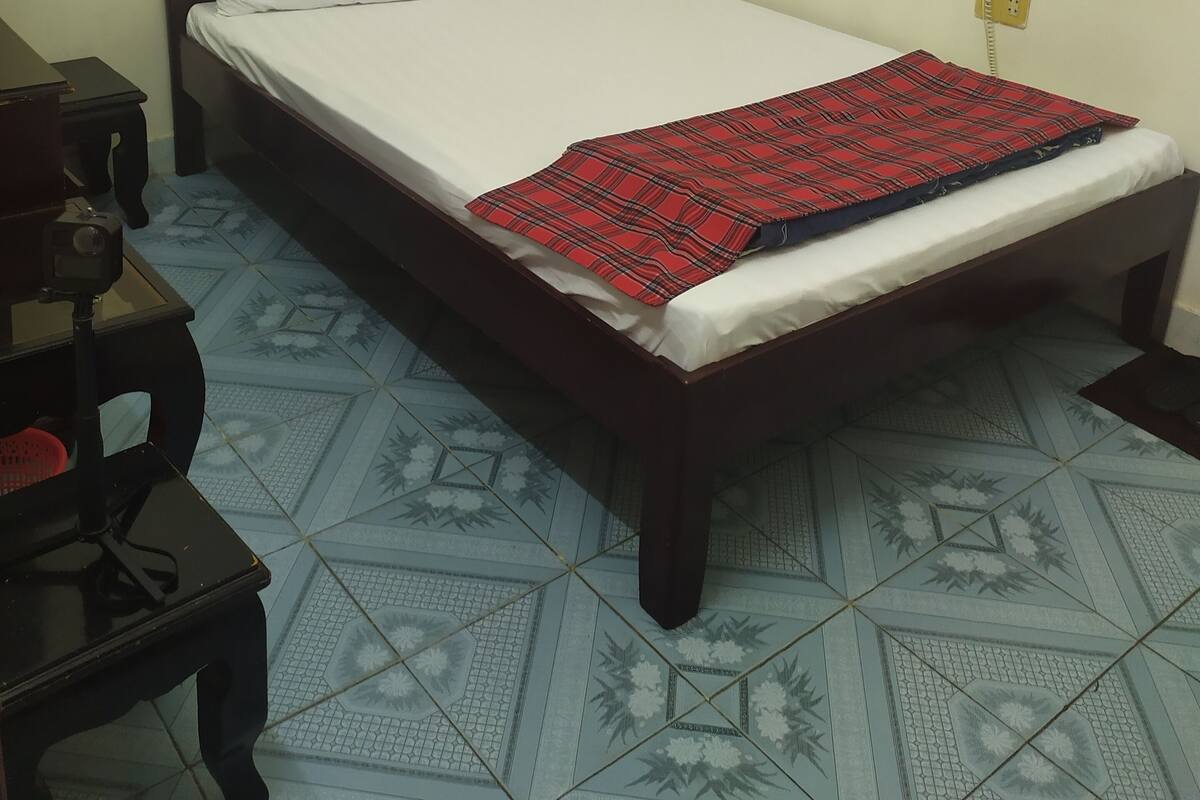 A single bed with white bedding and a red plaid blanket, a wooden nightstand, and a wall socket.