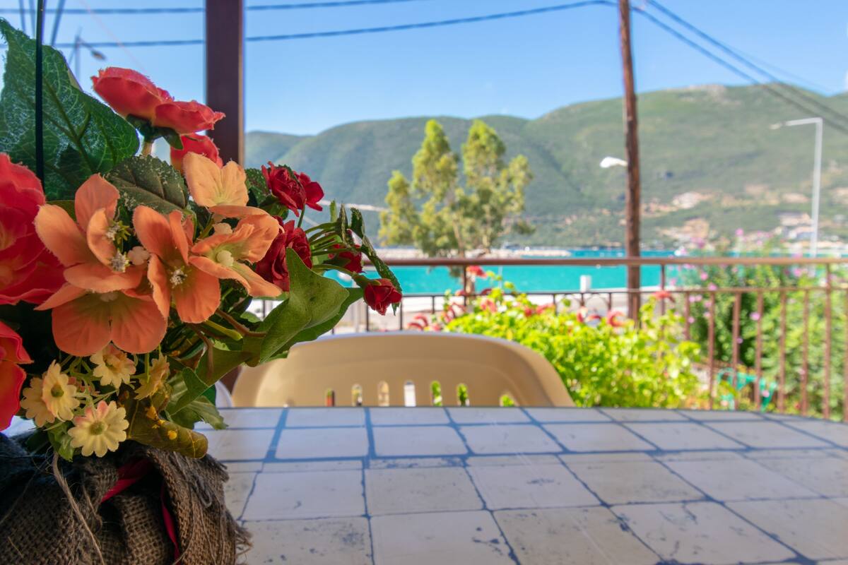 A table with a bouquet of flowers, a chair, and a view of a coastal area with mountains in the background.