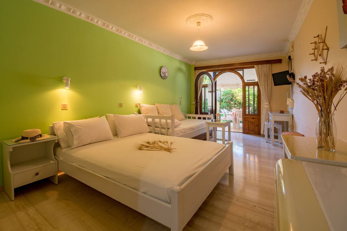 A bedroom with a large bed, bedside tables, a desk, and a chair. There is a window with curtains, a clock on the wall, and a vase with dried flowers on the desk.
