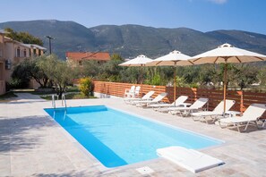 A swimming pool with lounge chairs and umbrellas, surrounded by a wooden fence and trees, with mountains in the background.