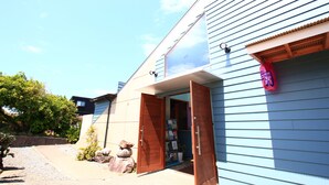A residential building with a wooden door open, revealing a small porch area with a mailbox and a garden with rocks and plants. - Rest and Lodge Syou (Amami)