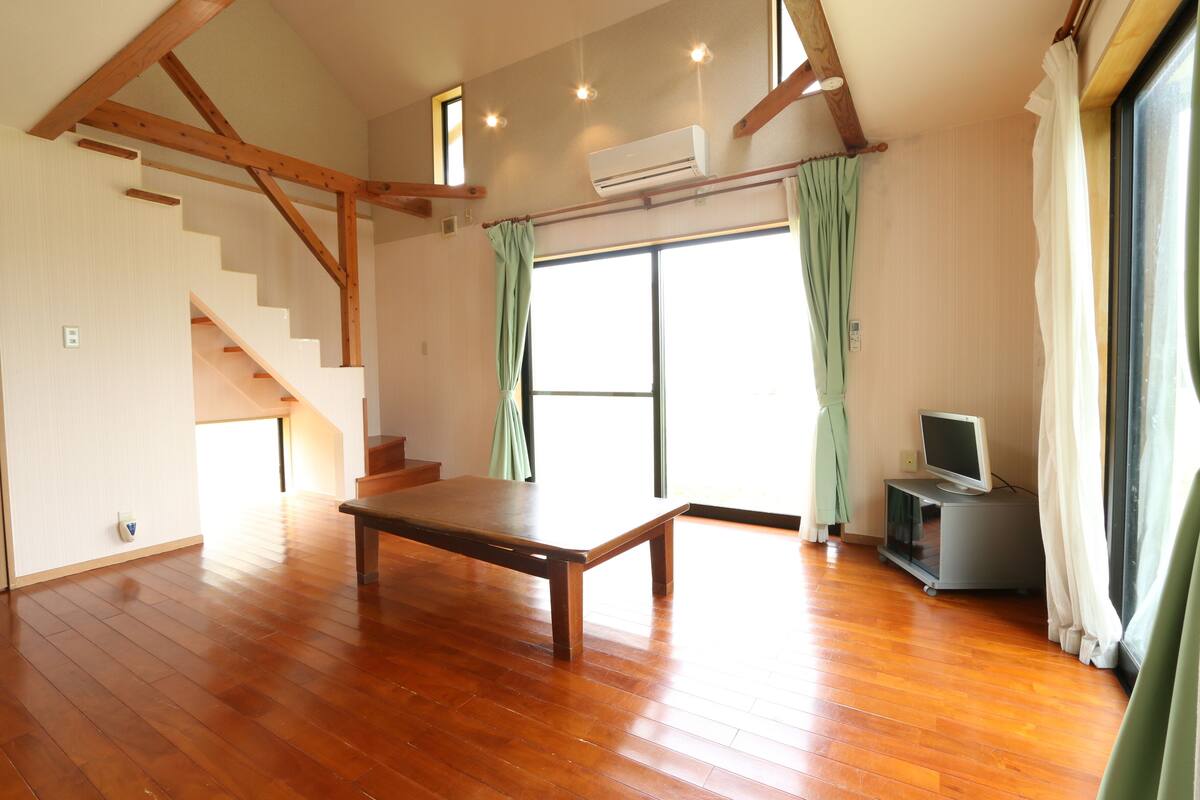 A spacious room with wooden flooring, a staircase, a flat-screen TV, and a large window with curtains.