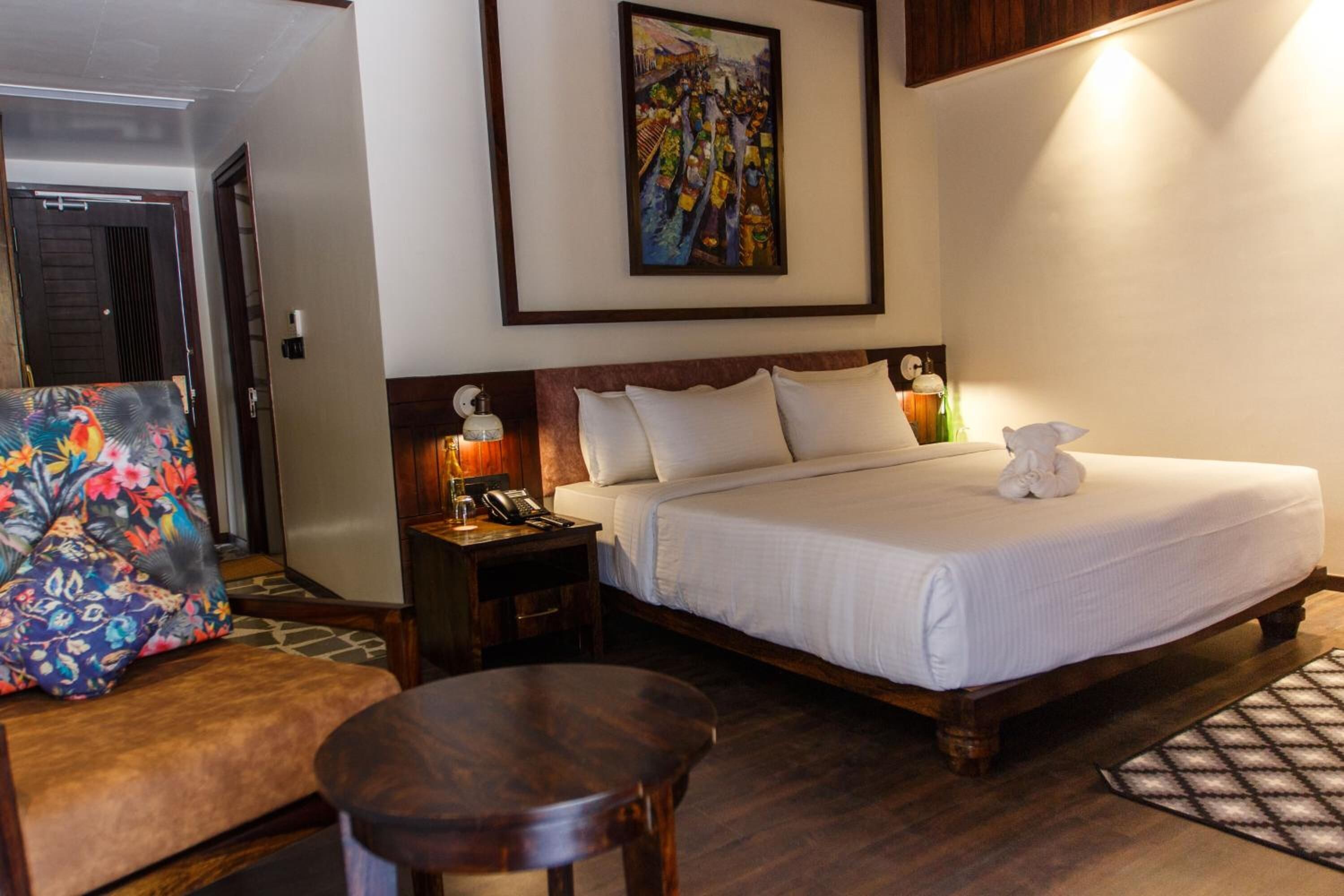 Una habitació d'hotel amb un llit, una taula de nit amb una làmpada, una cadira, un quadre a la paret i un terra de fusta.
