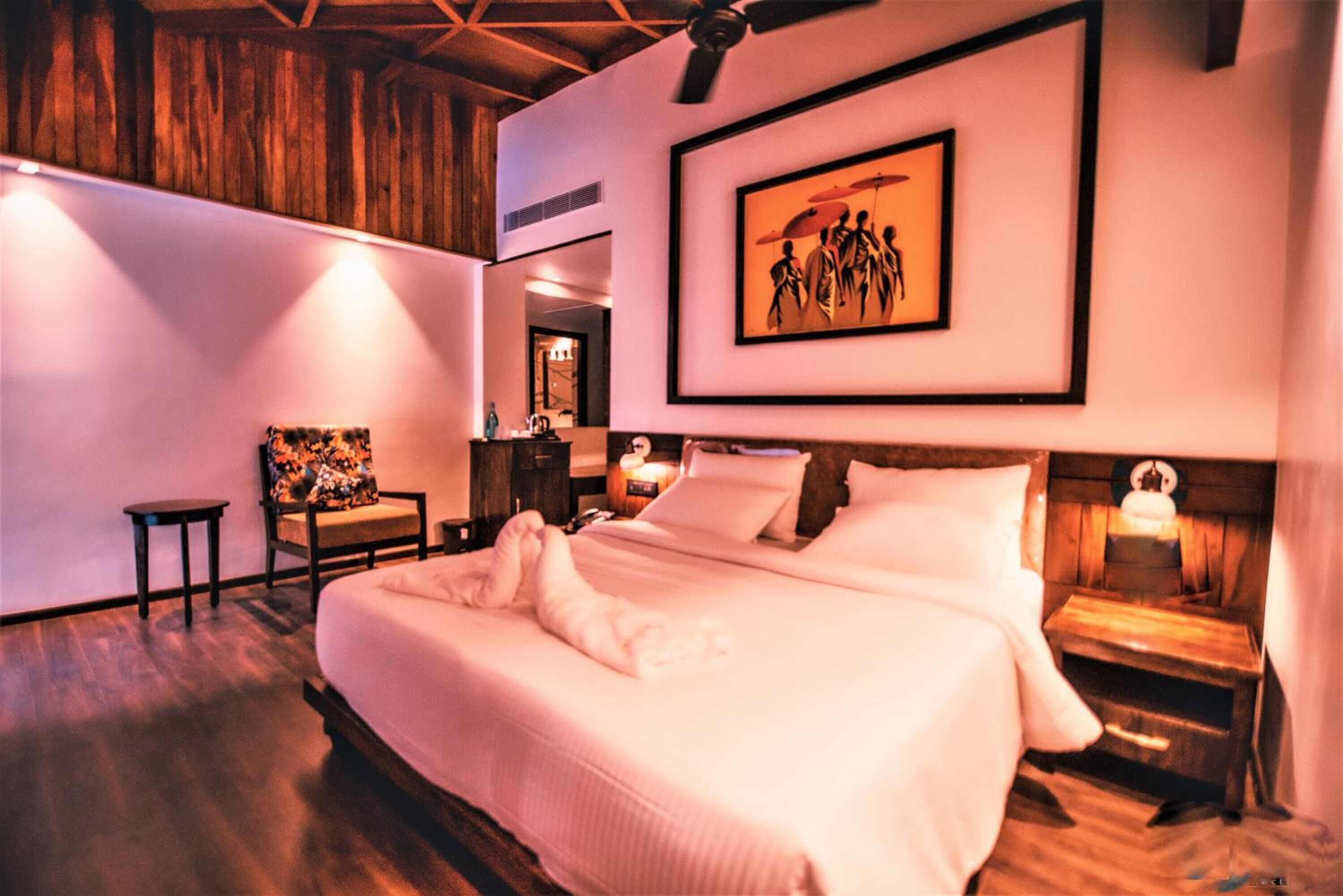 A neatly made bed with white linens, a wooden headboard, and a nightstand with a lamp. A framed artwork hangs above the bed, and a chair is positioned against the wall.