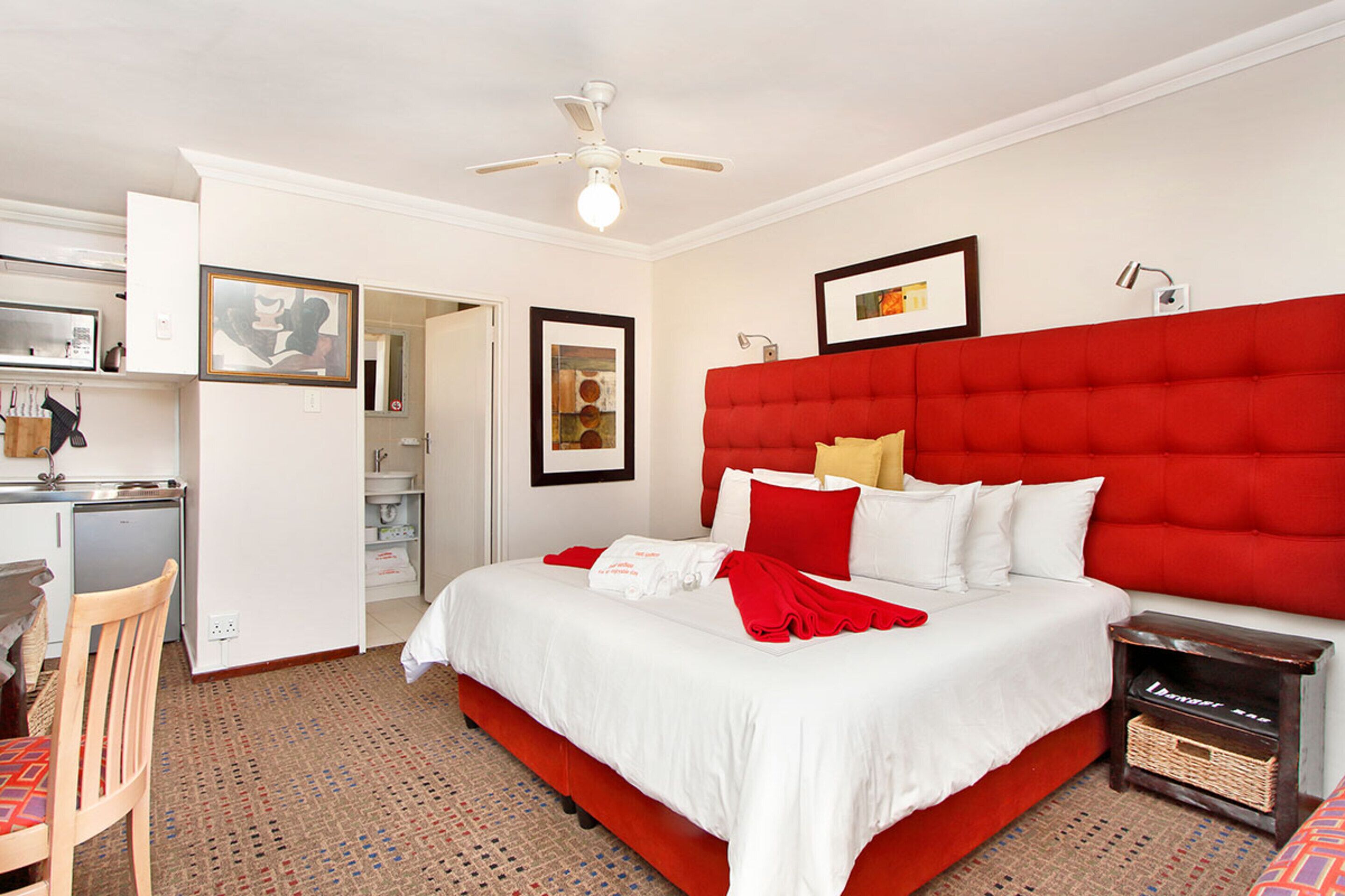 A hotel room with a large bed, a desk with a chair, a small kitchenette, and a ceiling fan.