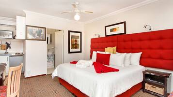 A hotel room with a large bed, a desk with a chair, a small kitchenette, and a ceiling fan.