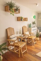 A room with wicker chairs, a wooden shelf with plants, a green wall light, and a white cabinet.