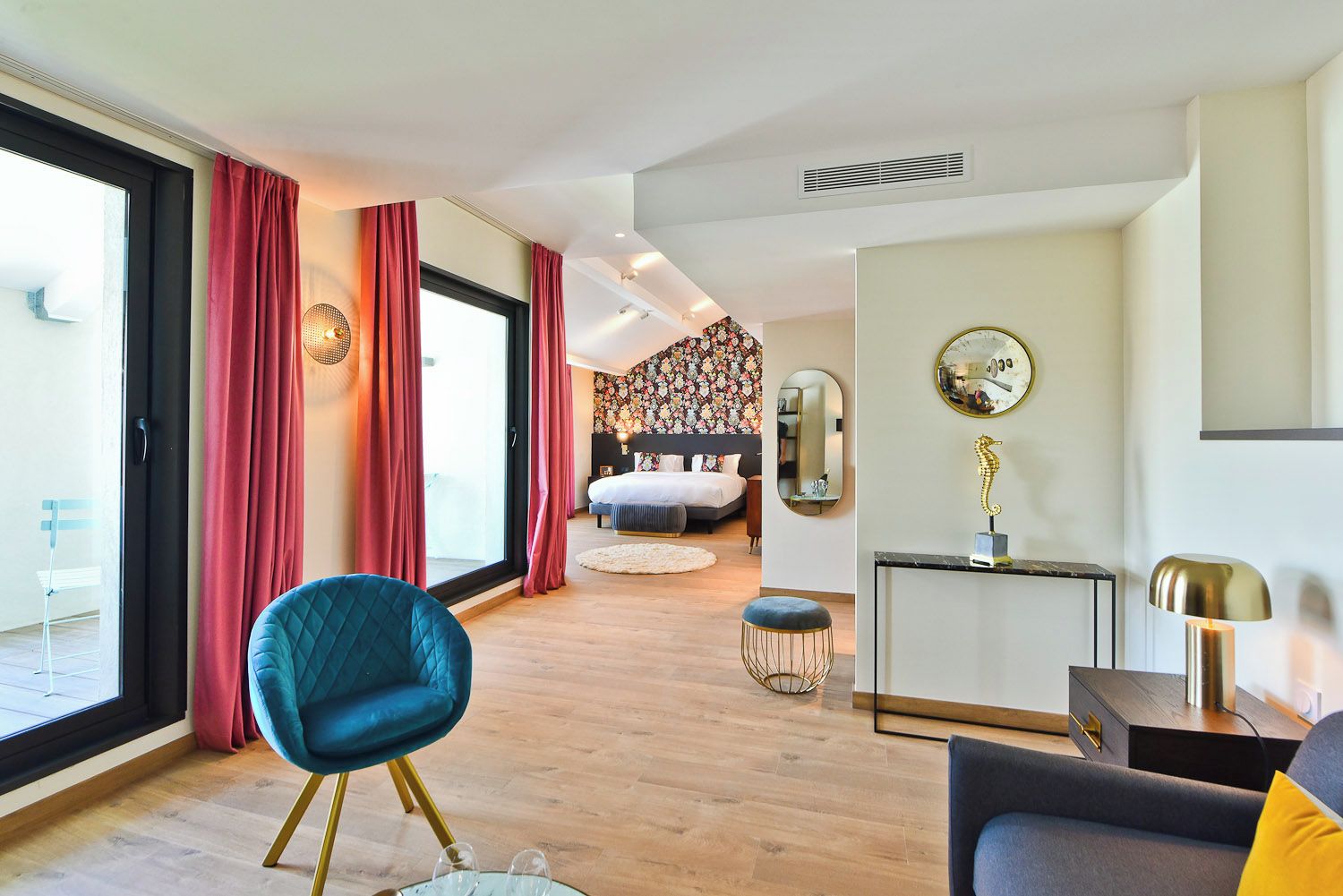 Une chambre d’hôtel moderne avec un grand lit, un coin salon comprenant un fauteuil bleu, une petite table ronde et un bureau avec une lampe.
