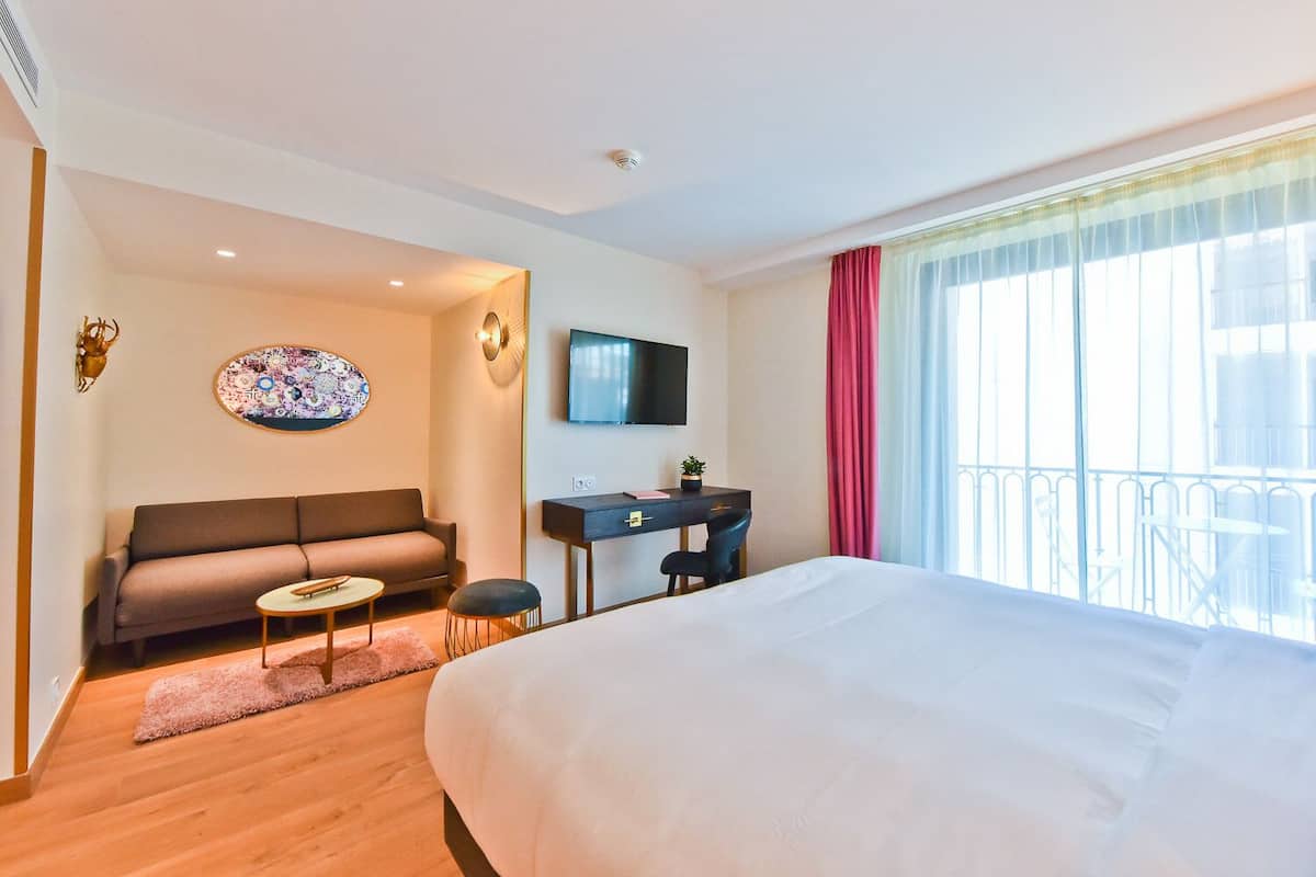 Habitación de hotel con cama, sofá, escritorio, televisor y balcón con barandilla.
