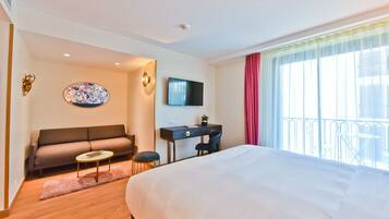A hotel room with a bed, a sofa, a desk, a TV, and a balcony with a railing.