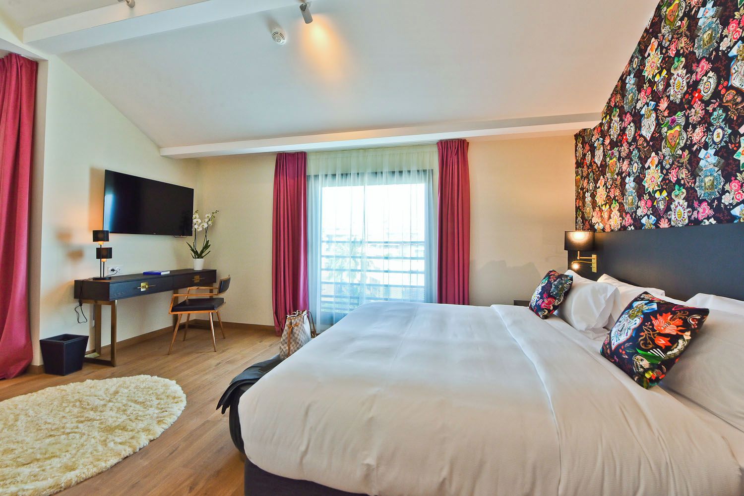 Quarto de hotel com uma cama grande, uma escrivaninha com televisão, uma janela com cortinas e uma tapeçaria colorida na parede.